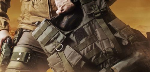 Photo of a soldier in military outfit holding a gun and bulletproof vest on orange desert background.